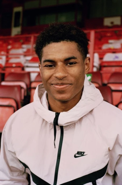 Close-up shot of a smiling, dark-skinned man in a light pink Nike hooded jacket, sitting in a red stadium seat. Sport and fashion.