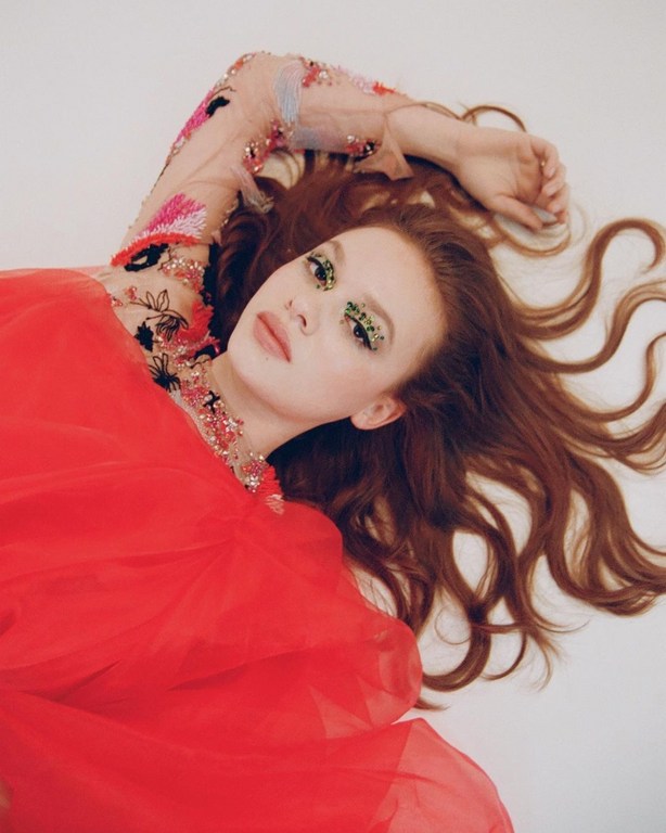 Model with long red hair and embellished sheer sleeve lying on a white background wearing a bright red dress.