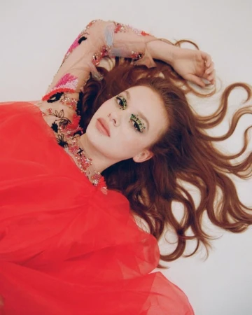 Model with long red hair and embellished sheer sleeve lying on a white background wearing a bright red dress.