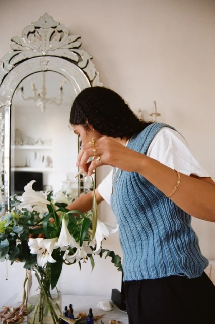 Adult woman arranges white lilies and greenery in a glass vase in an indoor home setting with a large mirror visible.