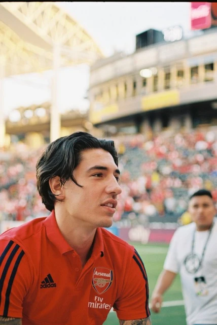 A man in a red Arsenal soccer jersey at an outdoor stadium, looking off-camera with a blurred crowd in the background.