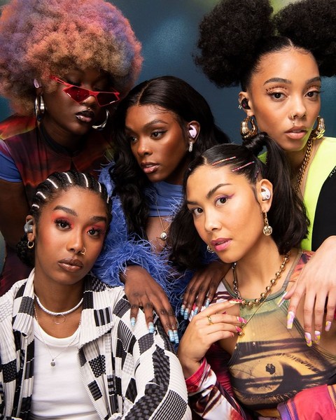Diverse group of five young women wearing earbuds, posing indoors with colorful clothing and jewelry, medium shot. Beauty, fashion, and tech.