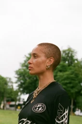Profile portrait of a woman with a buzz cut wearing a black graphic t-shirt and layered gold necklaces against an outdoor park background