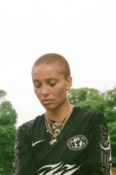 Close up portrait of a person with short buzzcut hair wearing a black long sleeve graphic top and layered gold chains in London