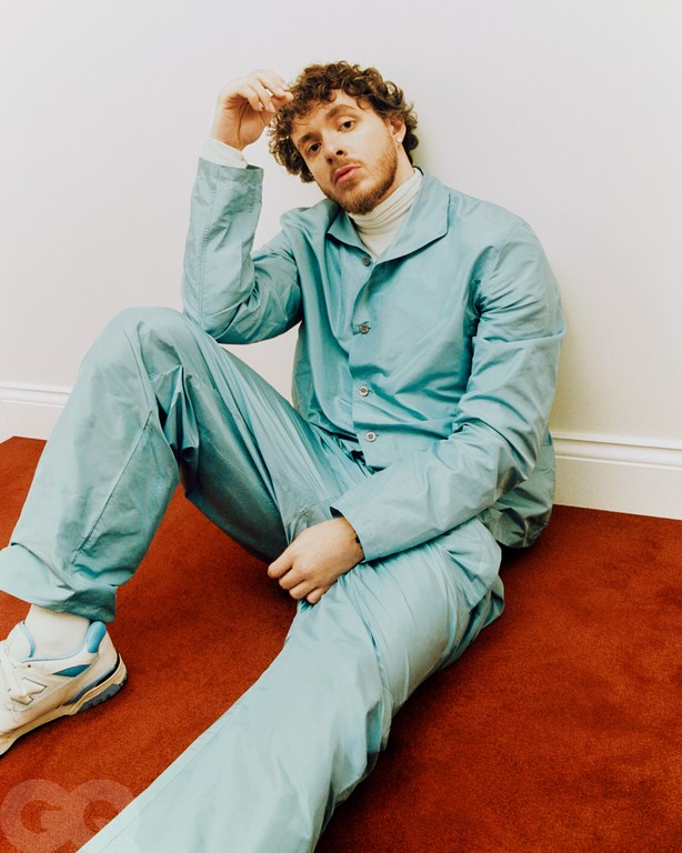 Adult male with curly hair, wearing a light blue jacket and pants, sitting on a red carpet against a white wall, casual pose.