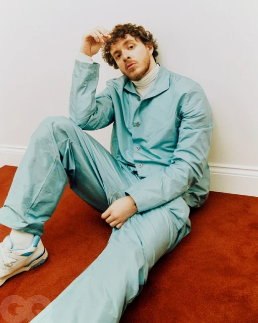 Adult male with curly hair, wearing a light blue jacket and pants, sitting on a red carpet against a white wall, casual pose.
