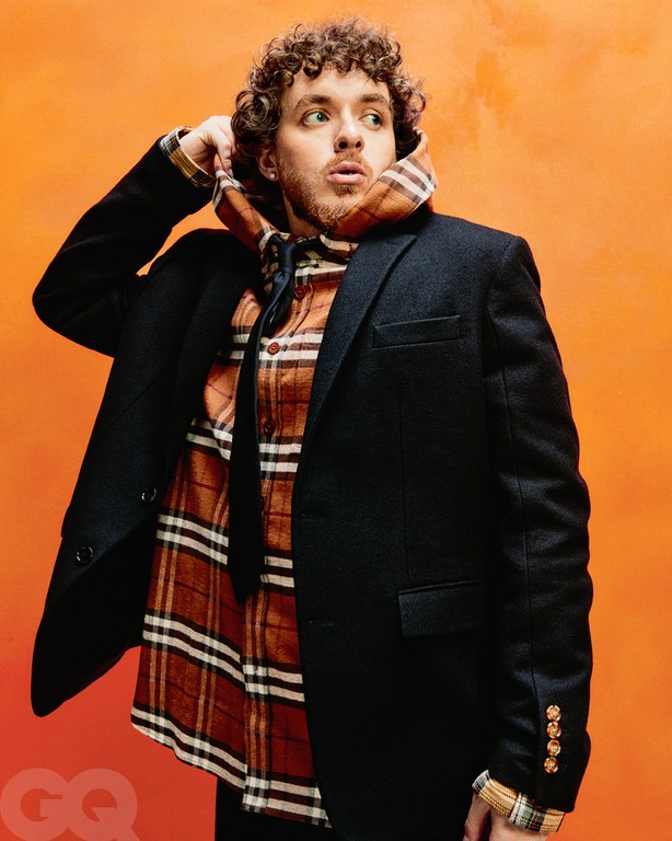 Jack Harlow posing in front of an orange background, wearing a black blazer, flannel shirt, and a patterned hooded scarf.