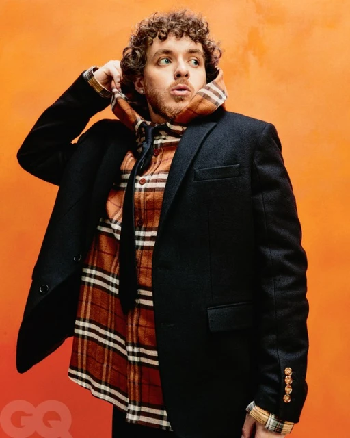 Jack Harlow posing in front of an orange background, wearing a black blazer, flannel shirt, and a patterned hooded scarf.