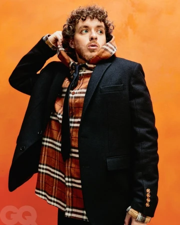 Jack Harlow posing in front of an orange background, wearing a black blazer, flannel shirt, and a patterned hooded scarf.