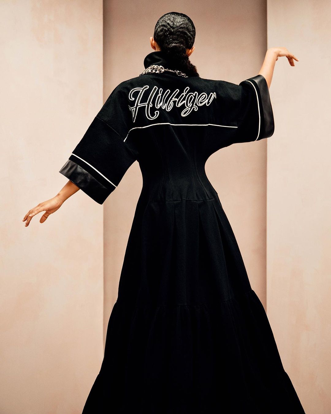 An adult female wearing a black, floor-length gown with 'Hilfiger' embroidered in white on the back poses in a photo studio.