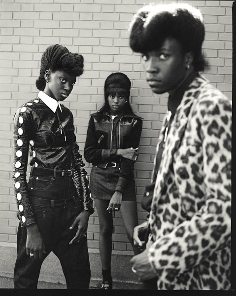 Black and white shot of three fashionably dressed young adults posing against a brick wall, showcasing stylish attire and confident expressions.