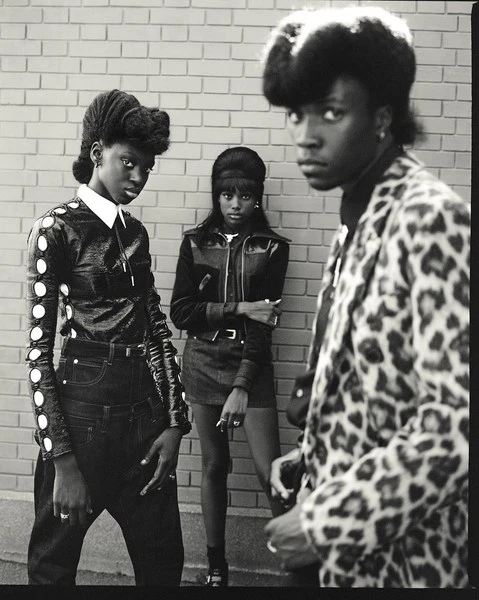 Black and white shot of three fashionably dressed young adults posing against a brick wall, showcasing stylish attire and confident expressions.