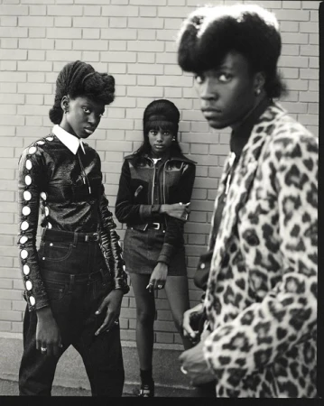 Black and white shot of three fashionably dressed young adults posing against a brick wall, showcasing stylish attire and confident expressions.