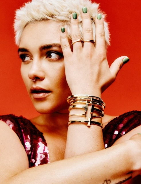 Close-up of a blonde woman wearing multiple gold bangle bracelets and rings, against a vibrant orange background in a studio.