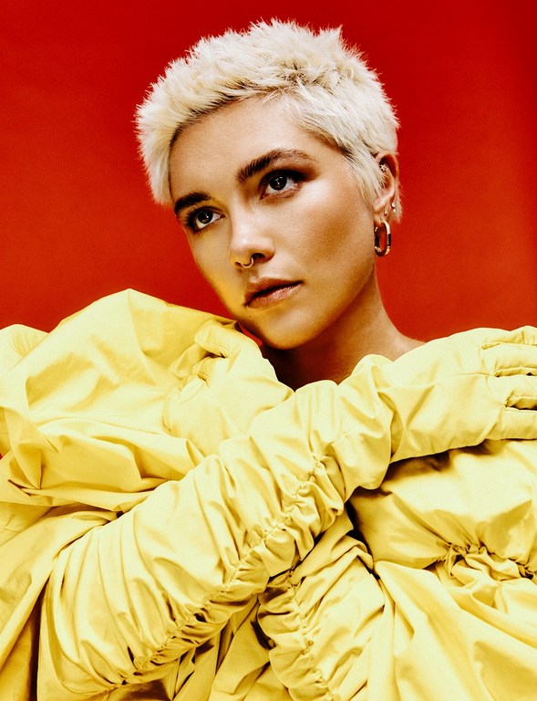 Portrait of Florence Pugh with short blonde hair wearing a bright yellow ruffled garment against a solid red backdrop