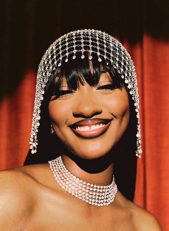 Portrait of a smiling, dark-skinned woman with a beaded headpiece and necklace against an orange curtain backdrop, close-up framing.