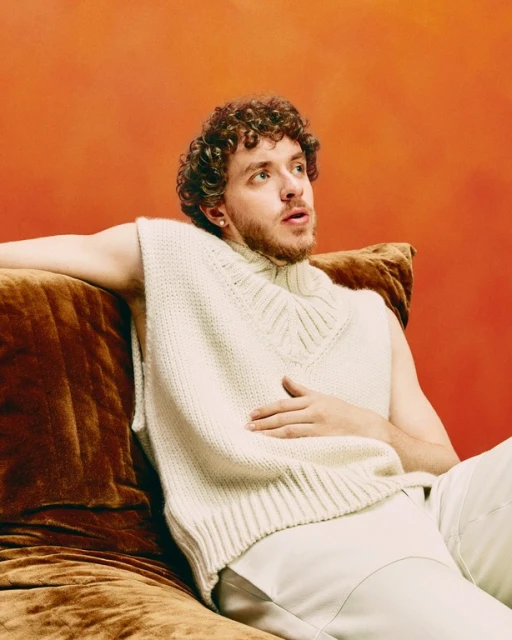 Portrait of curly haired adult male model in white knitted vest sitting on a brown velvet couch against a textured orange wall.