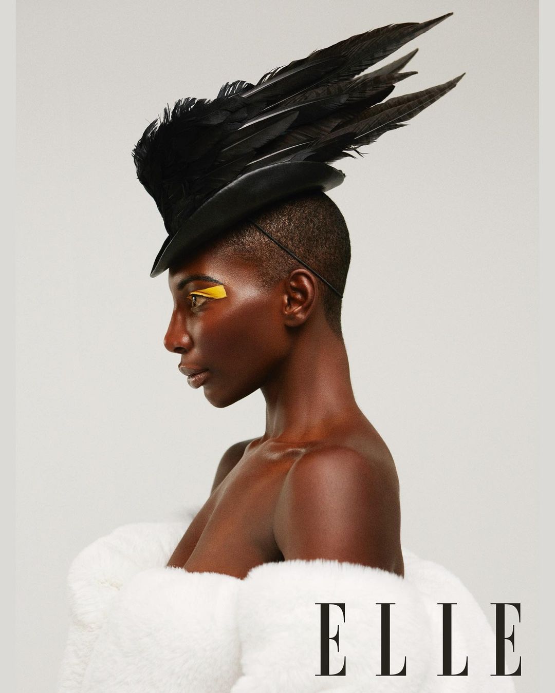 Profile portrait of a dark-skinned woman in a black feather hat, white fur coat, and gold makeup, shot in a bright photo studio.