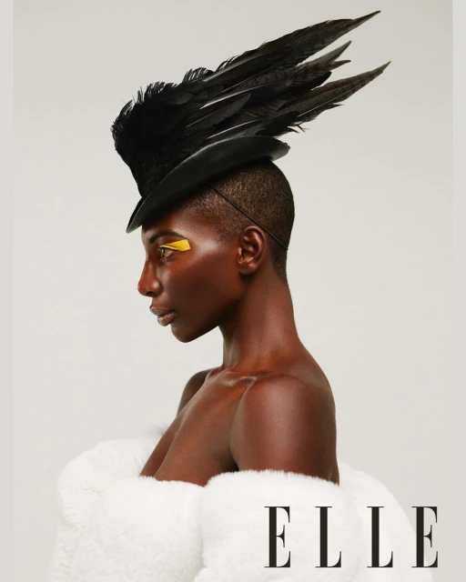 Profile portrait of a dark-skinned woman in a black feather hat, white fur coat, and gold makeup, shot in a bright photo studio.
