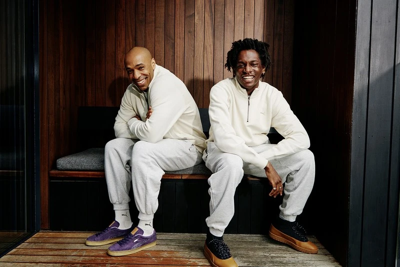 Two men sit side-by-side on a bench in an indoor setting, posing for a portrait. They wear matching casual outfits and branded Puma sneakers.