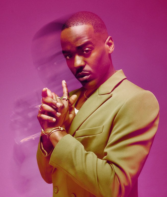 Headshot of a stylish Black male in a green blazer, posing confidently in a pink studio with fingers raised, showcasing jewelry.