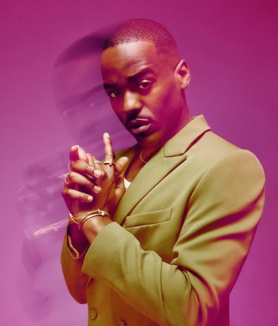 Headshot of a stylish Black male in a green blazer, posing confidently in a pink studio with fingers raised, showcasing jewelry.