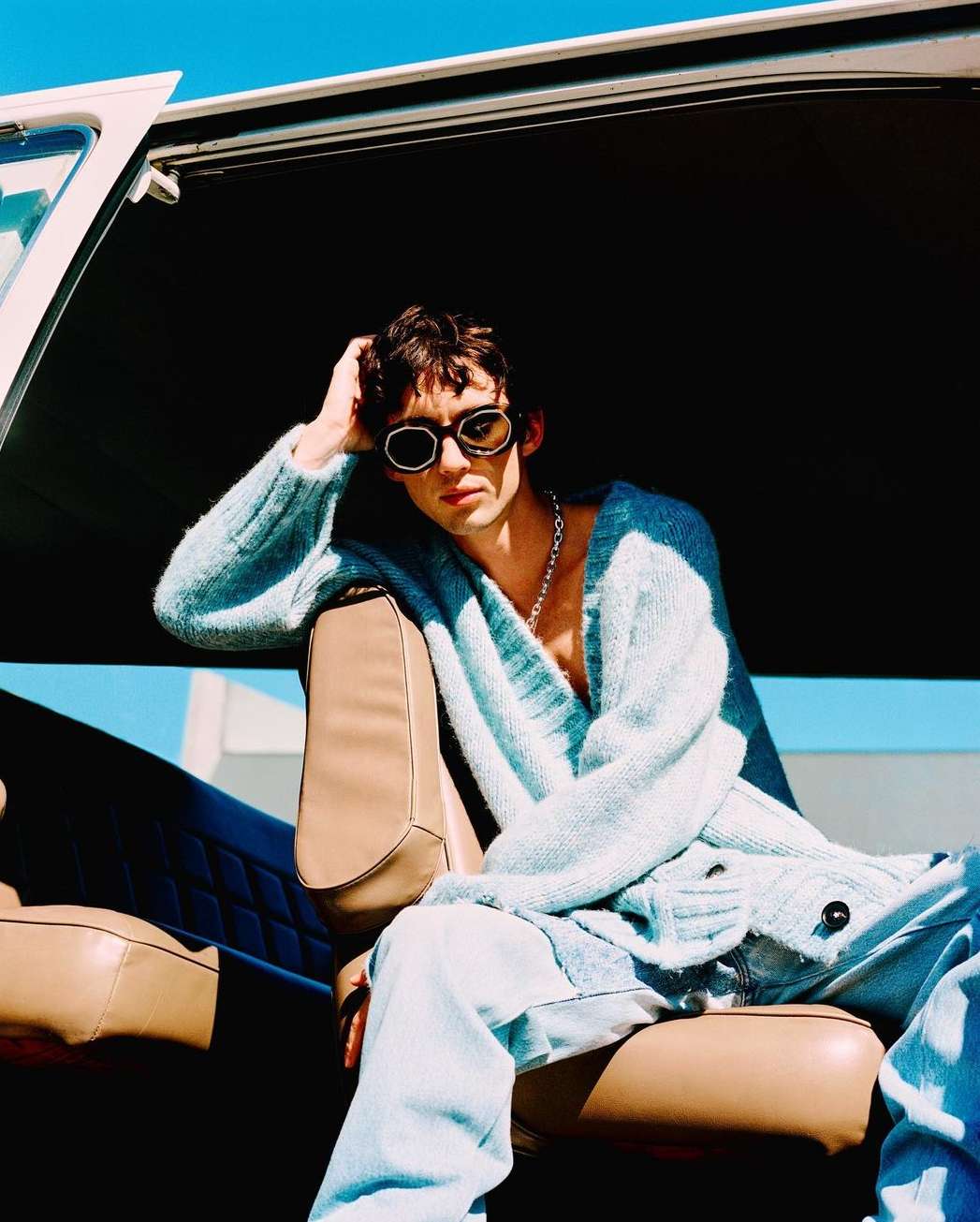 Adult male with sunglasses poses in a vehicle, wearing a blue cardigan and light blue jeans, with a white building in the background.