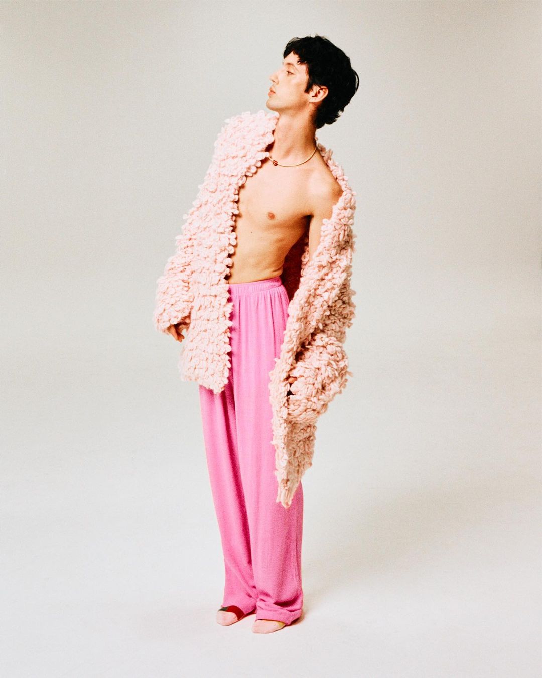A slender man with dark hair looks upward, wearing pink pants and a fuzzy pink jacket in a light studio, shot in portrait orientation.