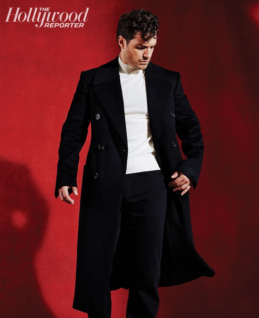 Henry Cavill wearing a black double-breasted overcoat and white turtleneck against a textured red background
