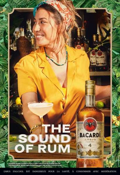 Woman in yellow shirt poses with Bacardi Añejo Cuatro rum bottle and cocktail against a backdrop of bar bottles and tropical leaves.