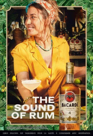 Woman in yellow shirt poses with Bacardi Añejo Cuatro rum bottle and cocktail against a backdrop of bar bottles and tropical leaves.