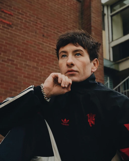Portrait of a young man with dark hair wearing an Adidas Manchester United jacket, posing against a brick wall backdrop.
