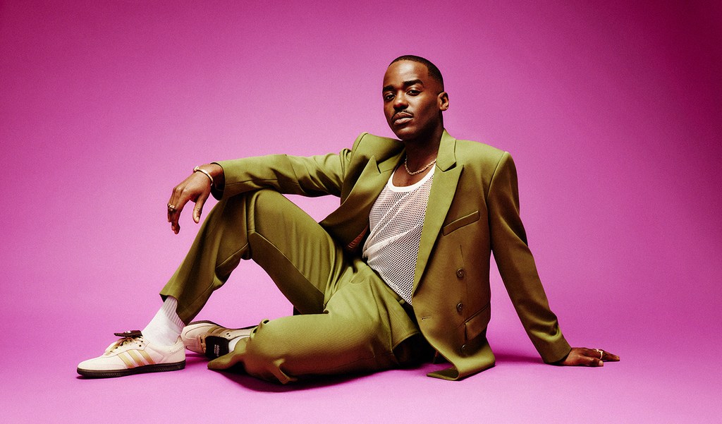Stylish man in an olive suit poses sitting on a pink background. He wears a white mesh shirt, sneakers, and gold jewelry.