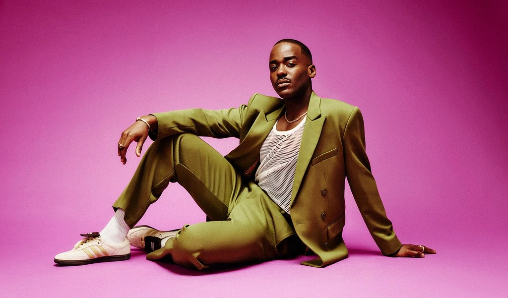 Stylish man in an olive suit poses sitting on a pink background. He wears a white mesh shirt, sneakers, and gold jewelry.