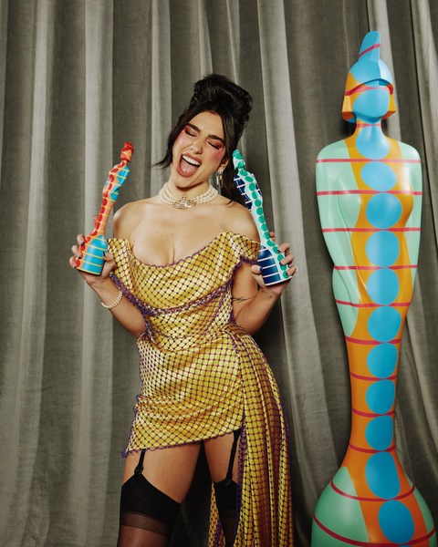 Singer Dua Lipa poses in a gold dress holding Brit Awards trophies in a photo studio setting with a statue beside her.