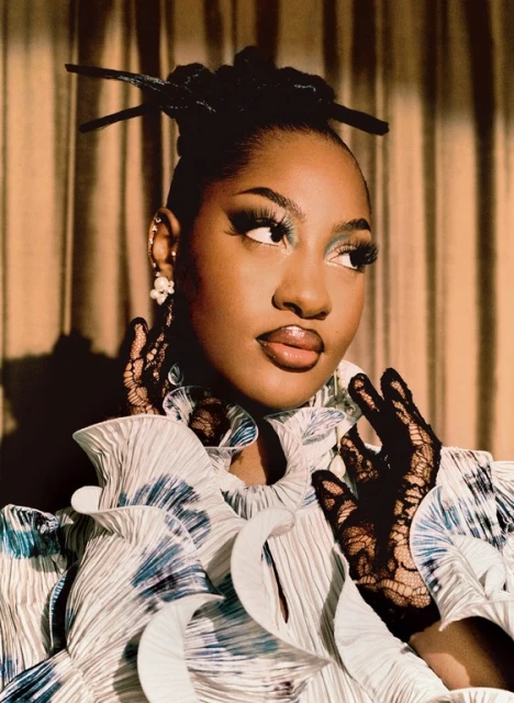 Portrait of a Black woman with elaborate updo, wearing a white ruffled dress with lace gloves against a gold curtain backdrop.