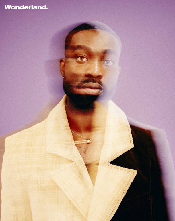 Portrait of a handsome, bearded Black man with short dark hair, wearing a two-tone blazer and pendant necklace against a purple backdrop.