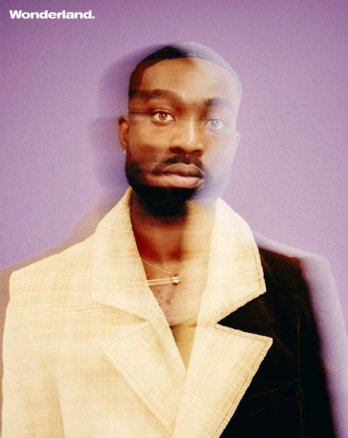 Portrait of a handsome, bearded Black man with short dark hair, wearing a two-tone blazer and pendant necklace against a purple backdrop.