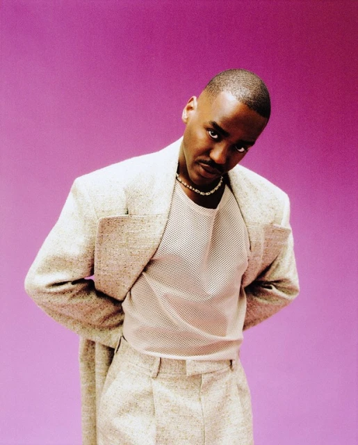 Portrait of a Black man with neutral expression in a beige suit jacket, wearing a silver chain, against a solid purple backdrop.