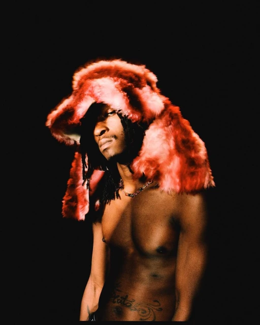 Portrait of a young adult with dreadlocks wearing a red and orange faux fur hat against a black background in a photo studio.