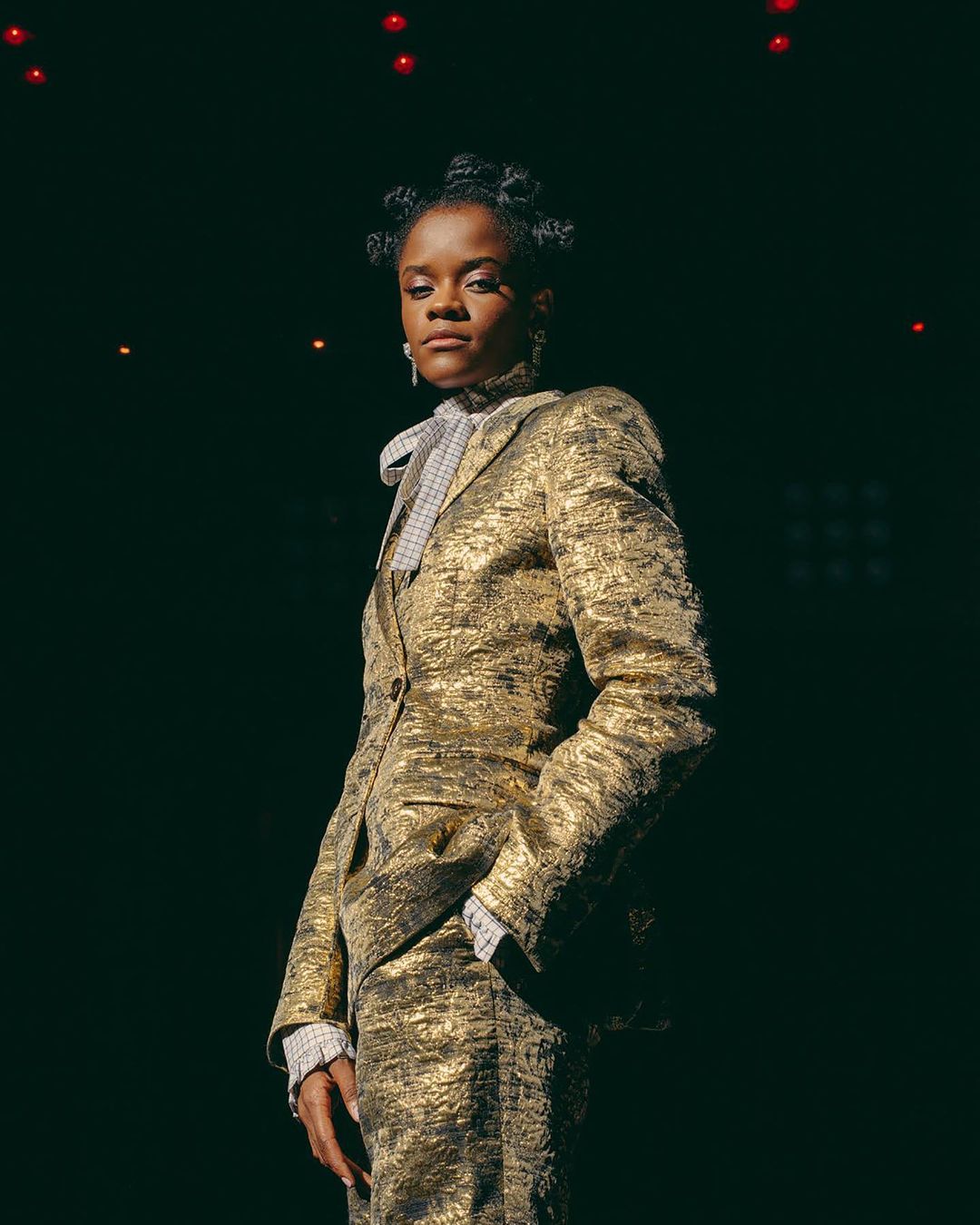 Headshot of Letitia Wright wearing a gold suit with hand in pocket against a black background with red accent lights.