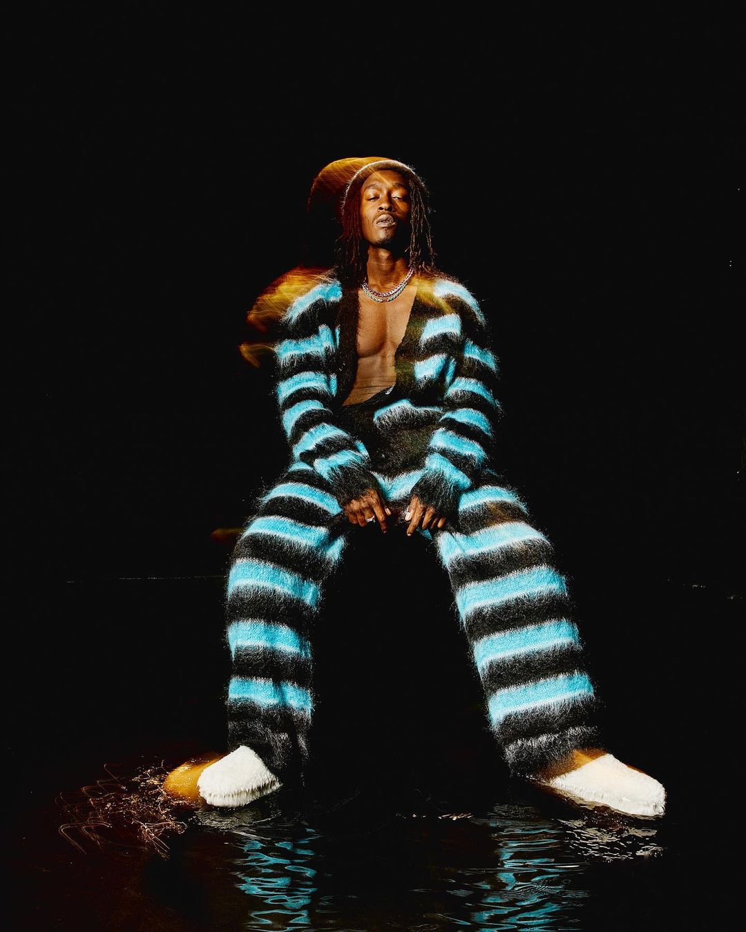 Man in striped loungewear sits in a dimly lit space with water reflections, wearing a silver chain and white slippers in a studio setting.