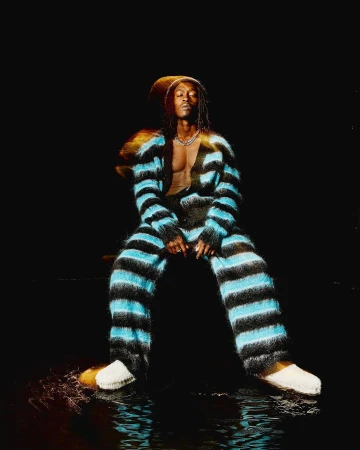 Man in striped loungewear sits in a dimly lit space with water reflections, wearing a silver chain and white slippers in a studio setting.