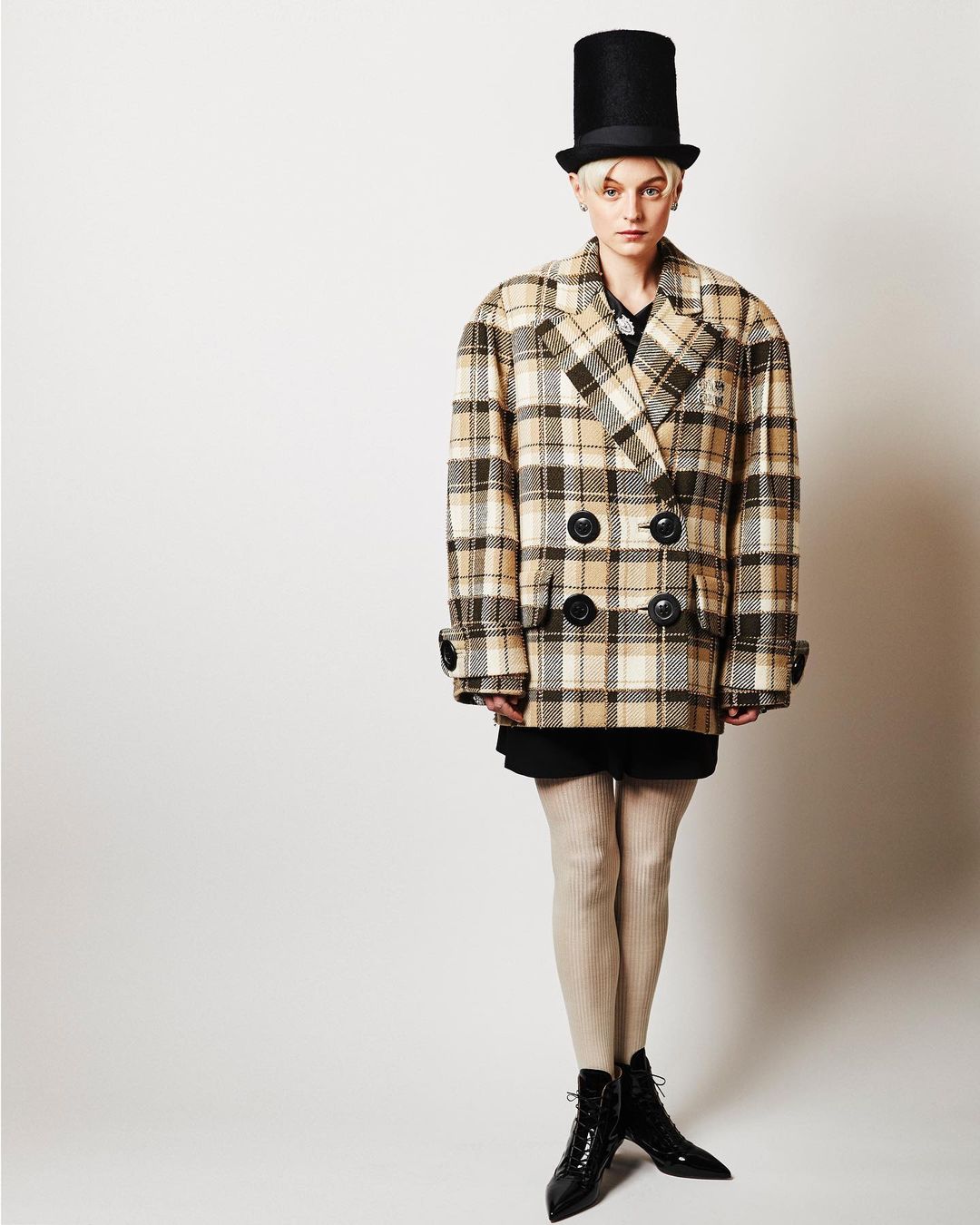 Woman in double-breasted plaid coat and shiny black heeled boots stands against a white backdrop, accessorized with a tall black hat.