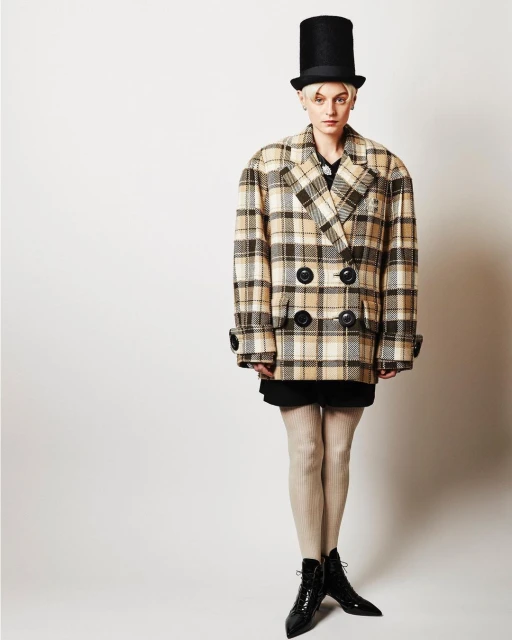 Woman in double-breasted plaid coat and shiny black heeled boots stands against a white backdrop, accessorized with a tall black hat.