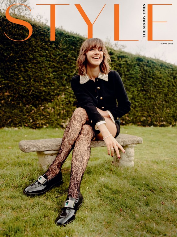 Magazine cover of a woman in a black jacket, tights, and loafers sitting on a bench in a grassy outdoor setting with a hedge backdrop.