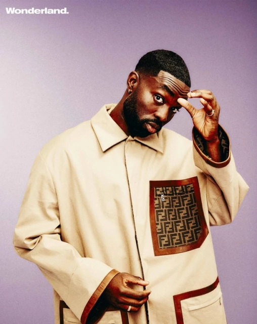 Close-up portrait of a black male model wearing a tan overcoat with patterned pockets on a light purple background in studio lighting.