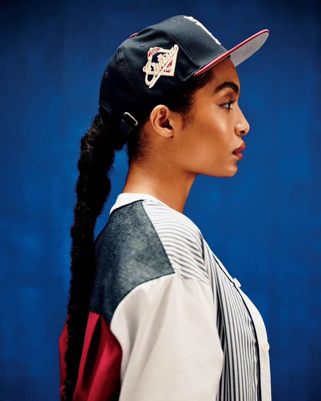 Side profile portrait of a young woman wearing a baseball cap and stylish color-blocked shirt against a solid blue backdrop.