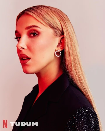 Portrait of Millie Bobby Brown wearing a black jacket and diamond hoop earrings against a neutral background