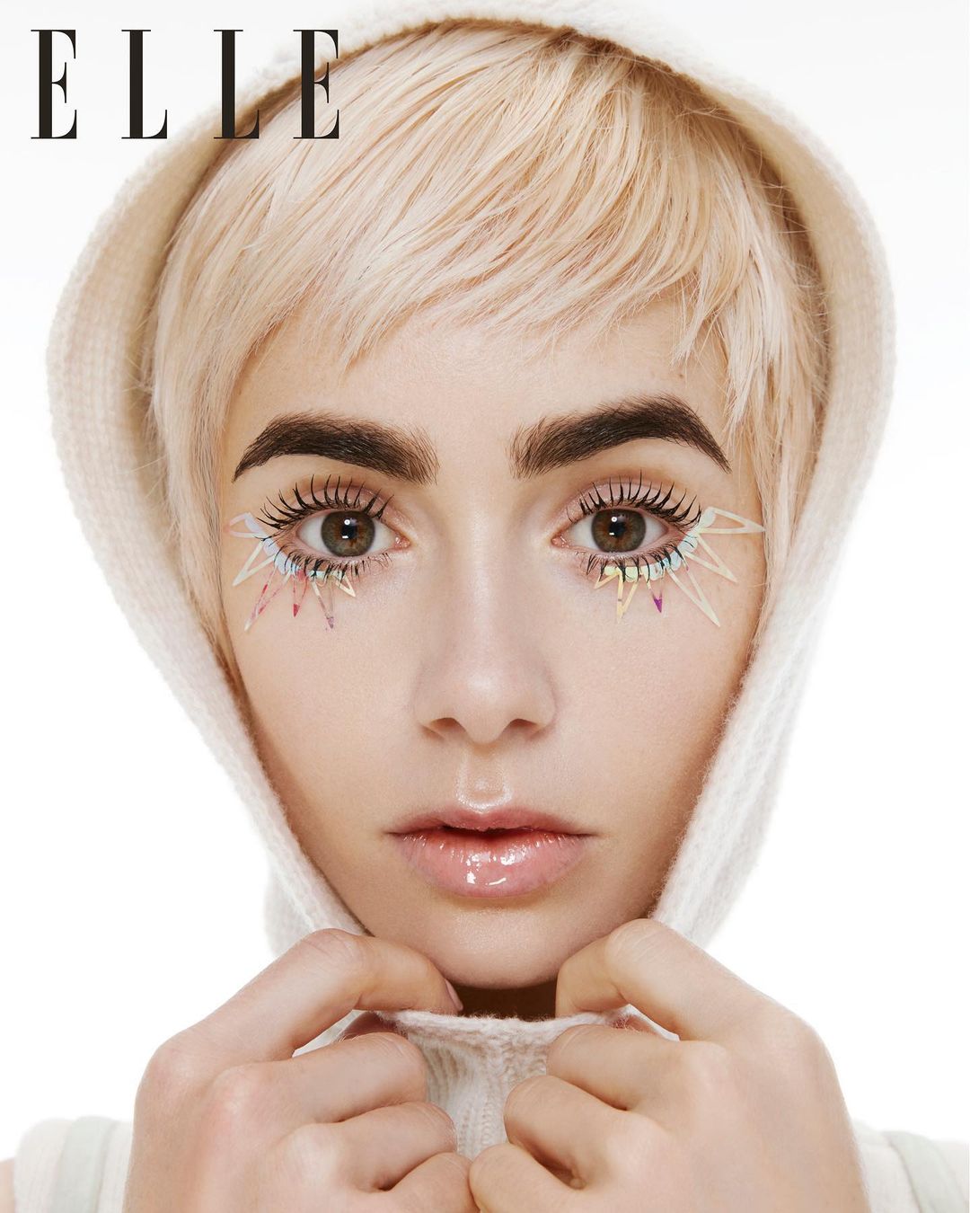 Close-up portrait of a woman with short blonde hair and hooded white top wearing colourful spiked lower eyelashes against white background.